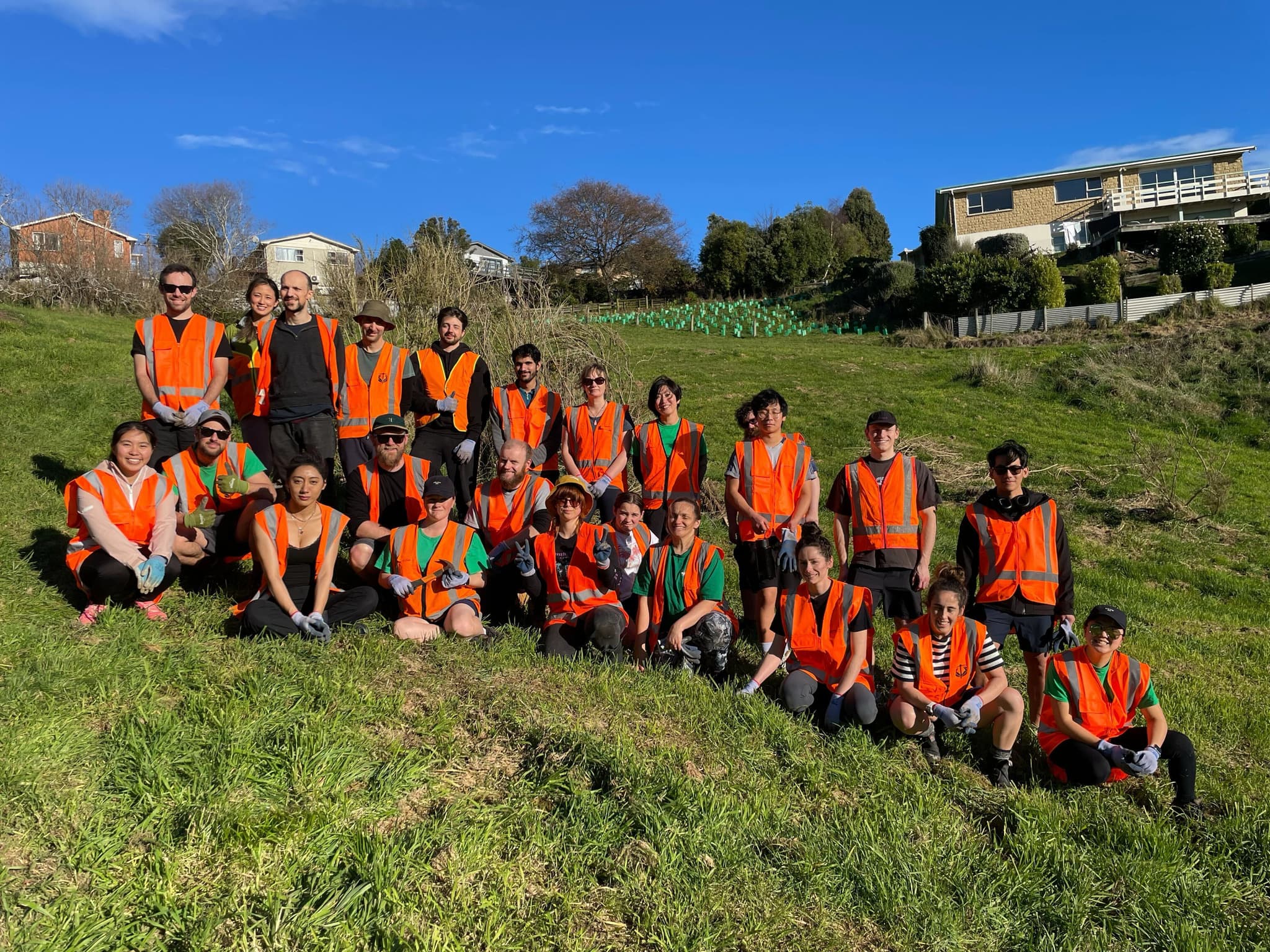 A large group in orange high-visibility vests and work gloves, posed in two rows on a grassy hillside, with young trees in guards and houses behind them under a blue sky.