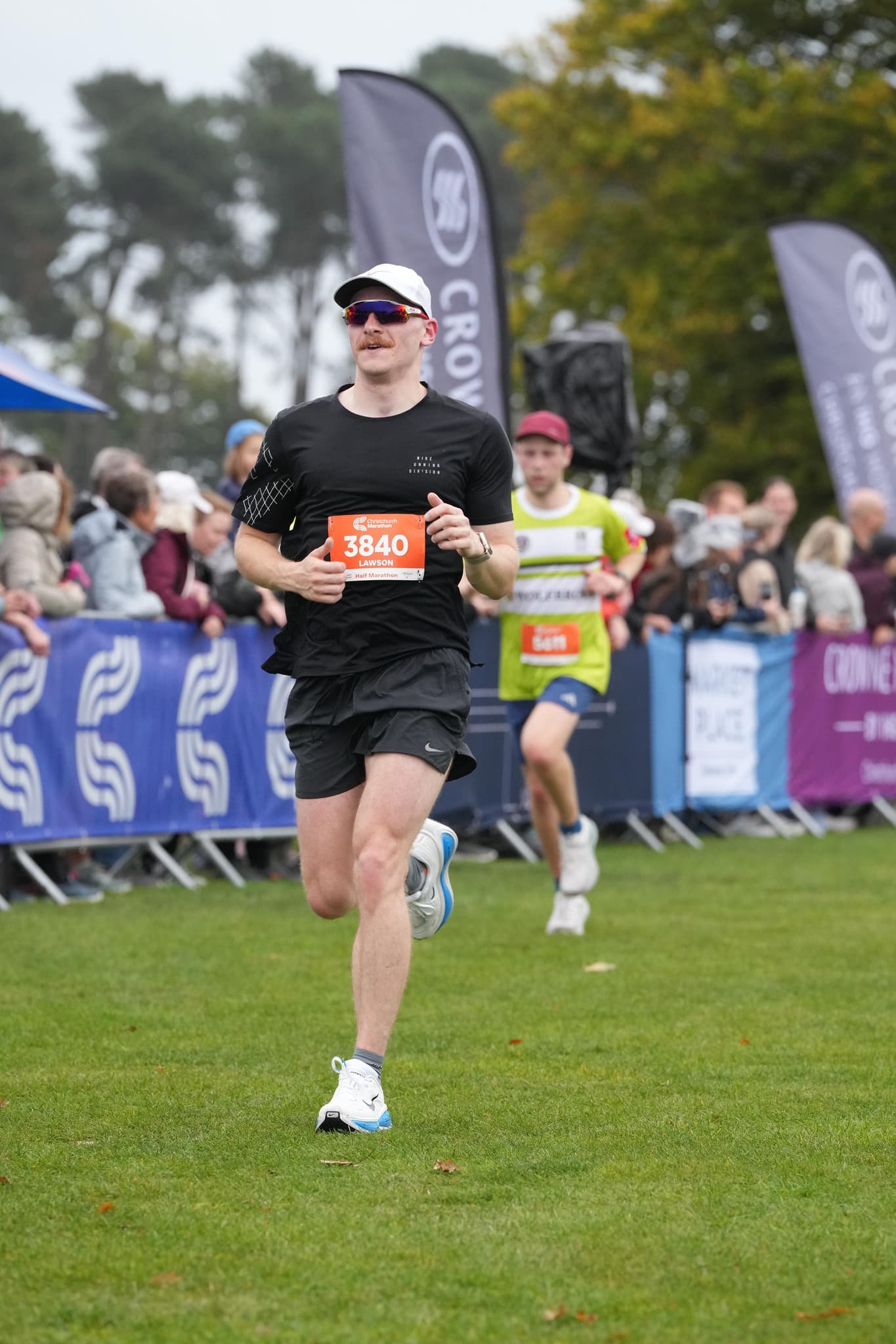 A runner in a white cap, sunglasses and black kit, wearing race number 3840, running on grass past a crowd behind barriers.