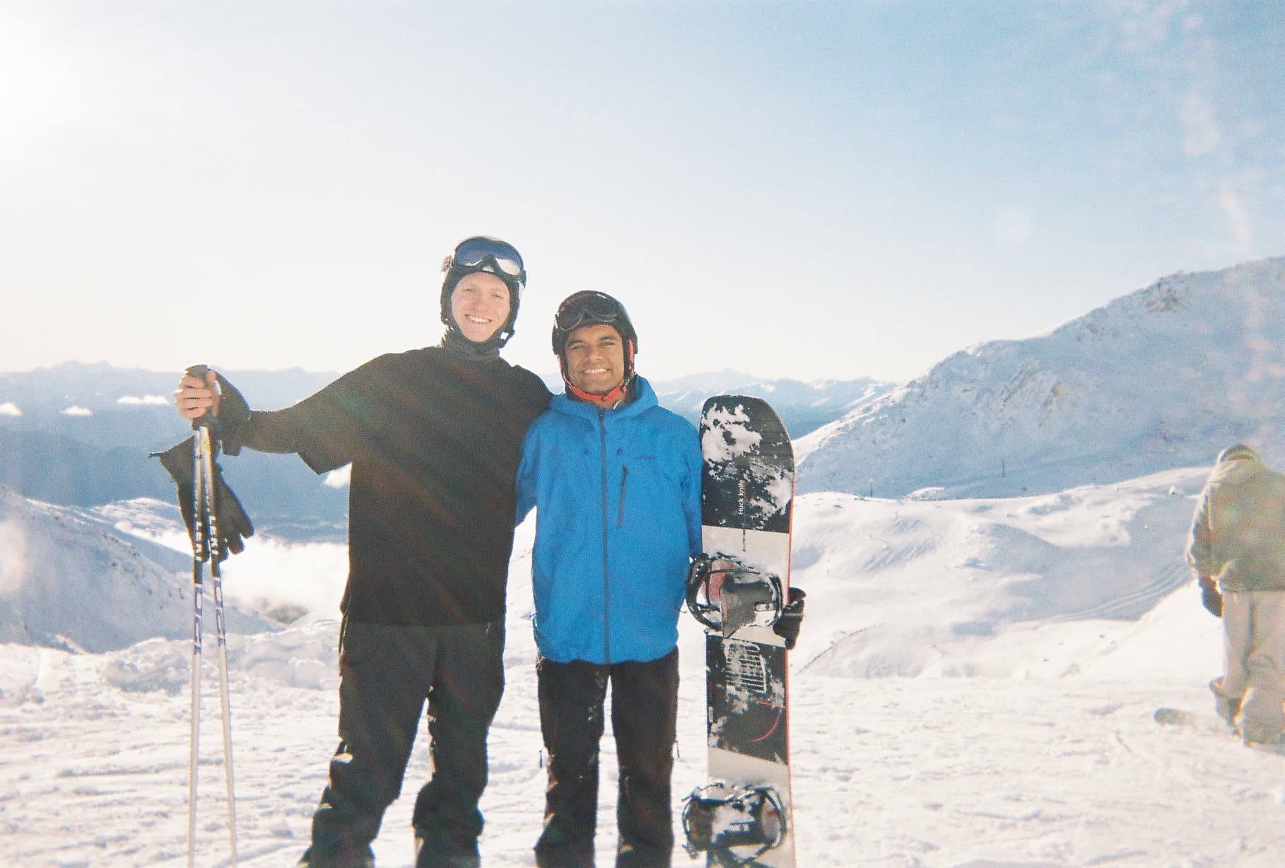 Two people on a snowy mountaintop under a bright sky, arms around each other: one in a black jacket holding ski poles, the other in a blue jacket holding a snowboard. Snow-covered ridges and low cloud fill the view behind them.
