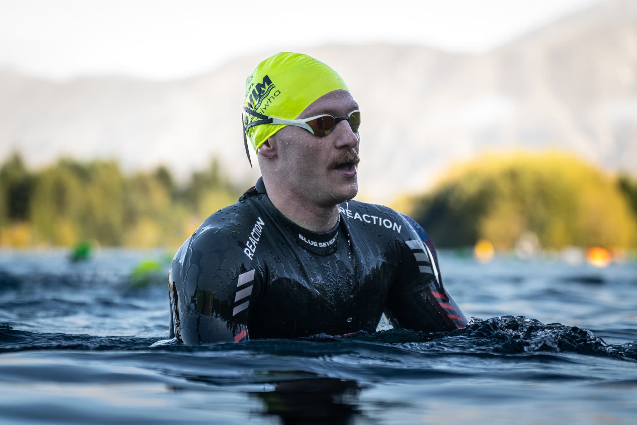 A swimmer in a yellow cap, mirrored goggles and a black wetsuit, shoulder-deep in a lake, with blurred trees and hills behind.