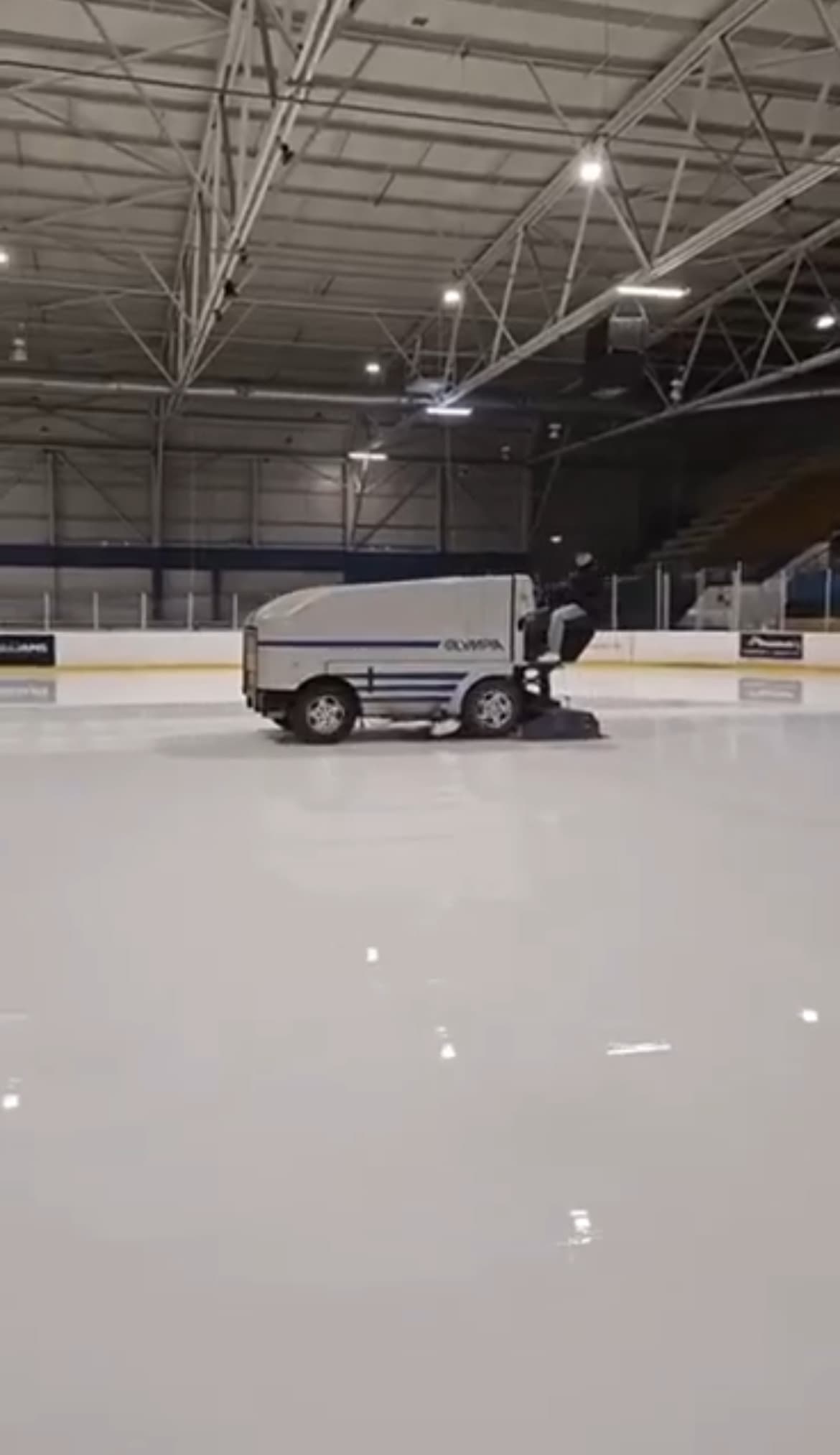 An ice resurfacer crossing an empty indoor rink under a steel-trussed roof.