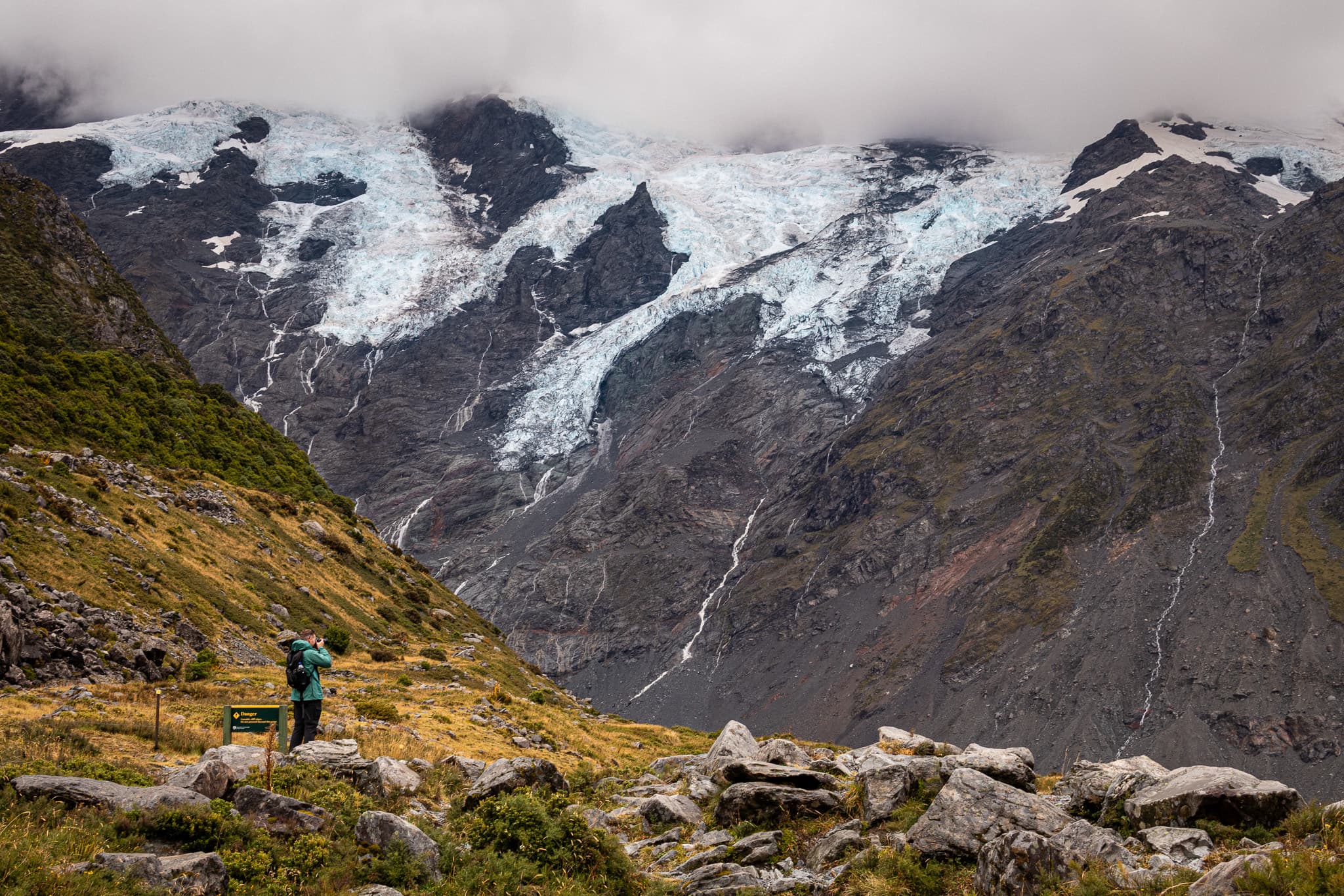 A person in a green jacket photographs glaciers spilling down a dark mountainside under low cloud, with meltwater streams running from the ice.