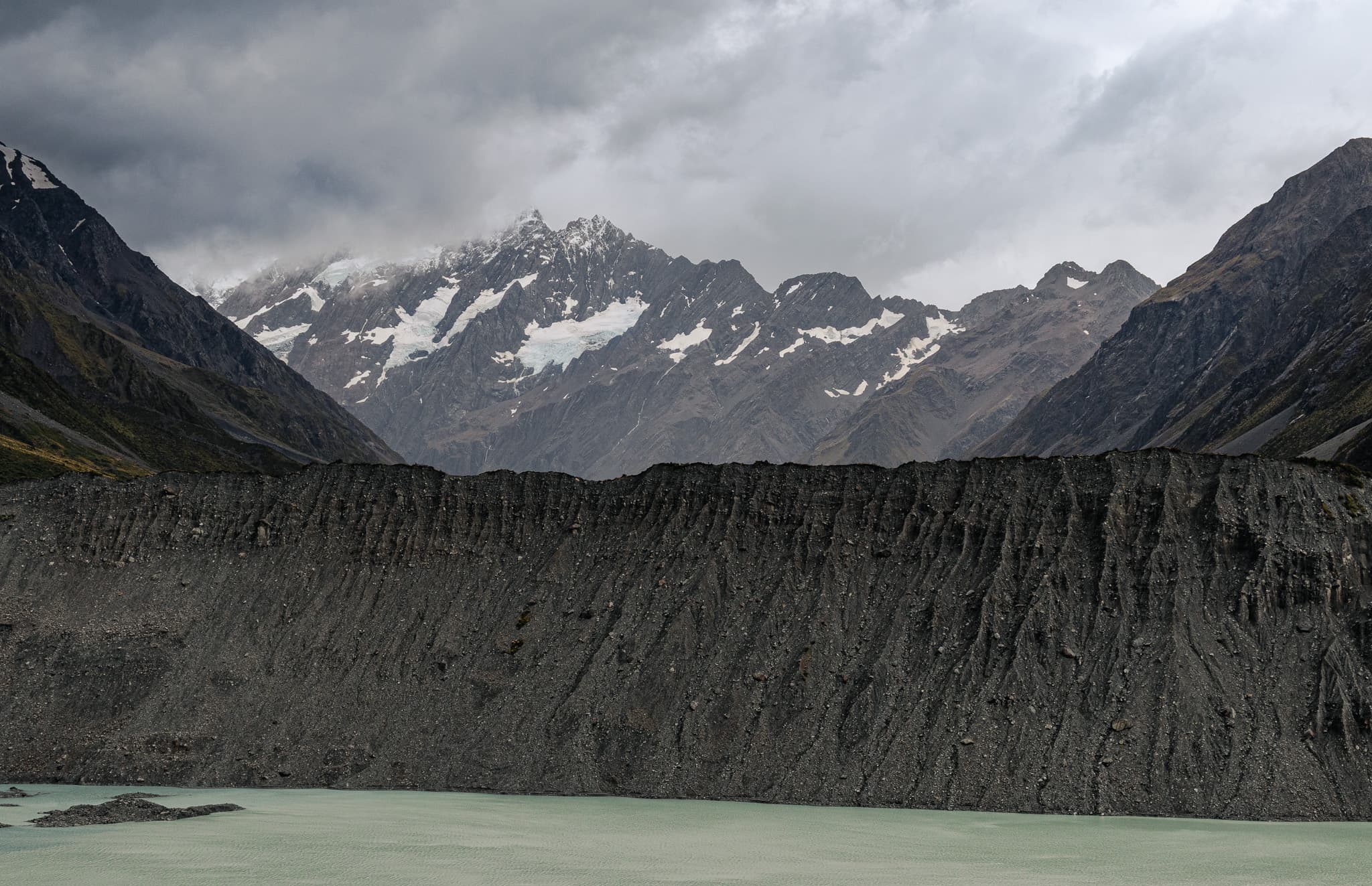 A steep grey wall of moraine above pale green water, with snow-streaked peaks under heavy cloud behind it.