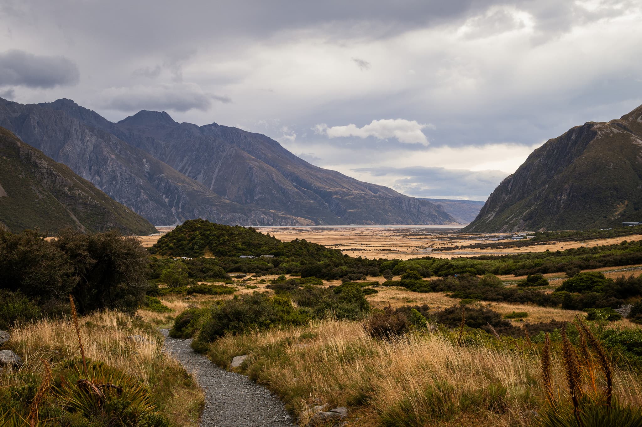 A gravel track through golden tussock and scrub, leading towards a wide valley between dark mountains under cloud.
