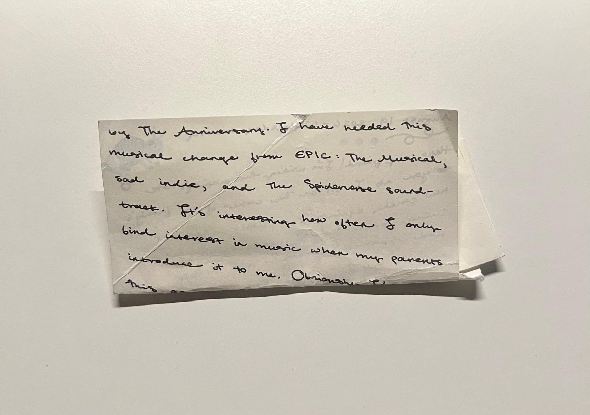 A small, creased slip of paper covered in neat handwriting, on a plain white surface. The lines that show are about music the writer has been listening to.