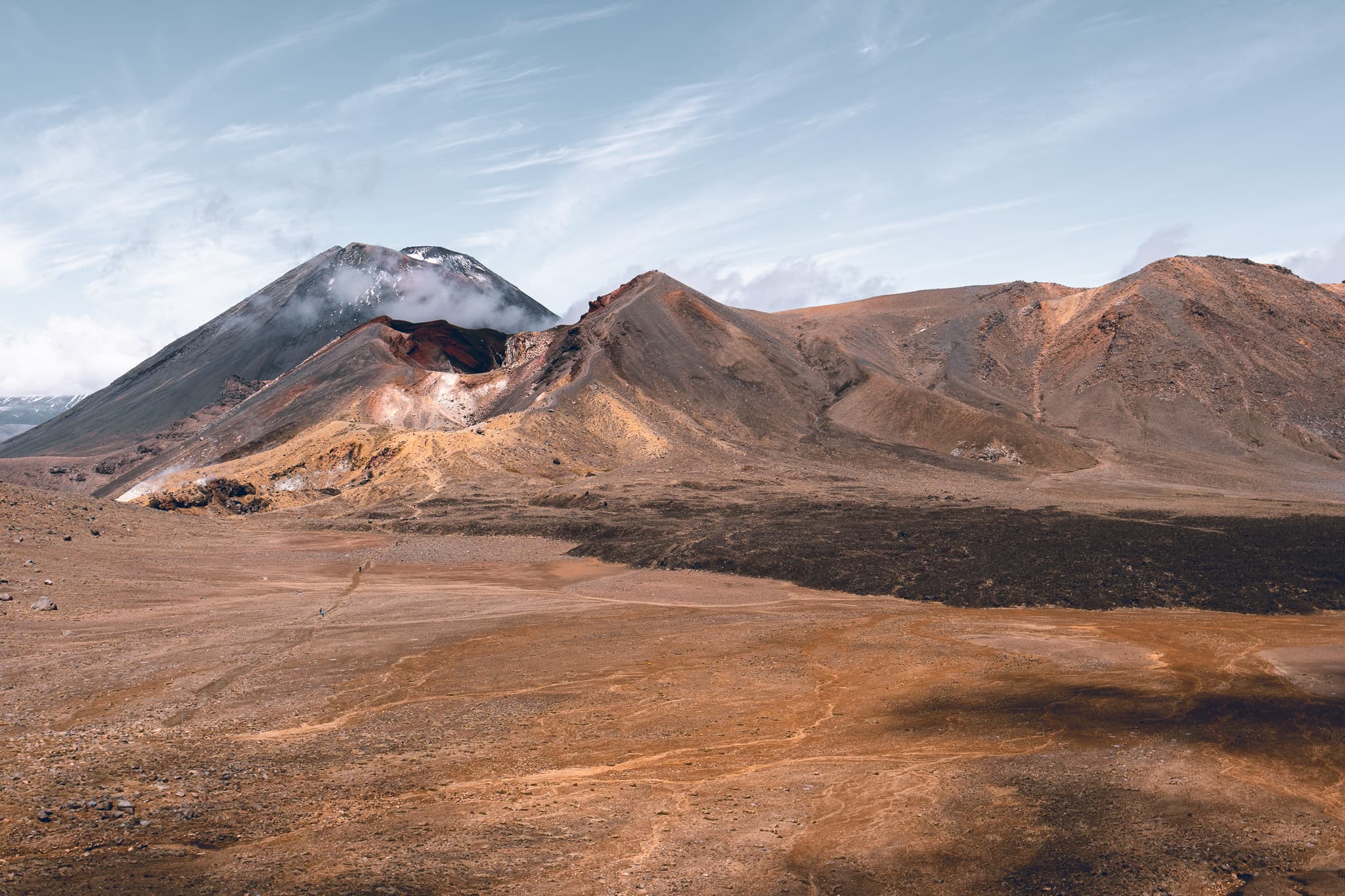 A snow-dusted volcanic cone half in cloud, above red and ochre craters and a wide, bare plain of volcanic ground.